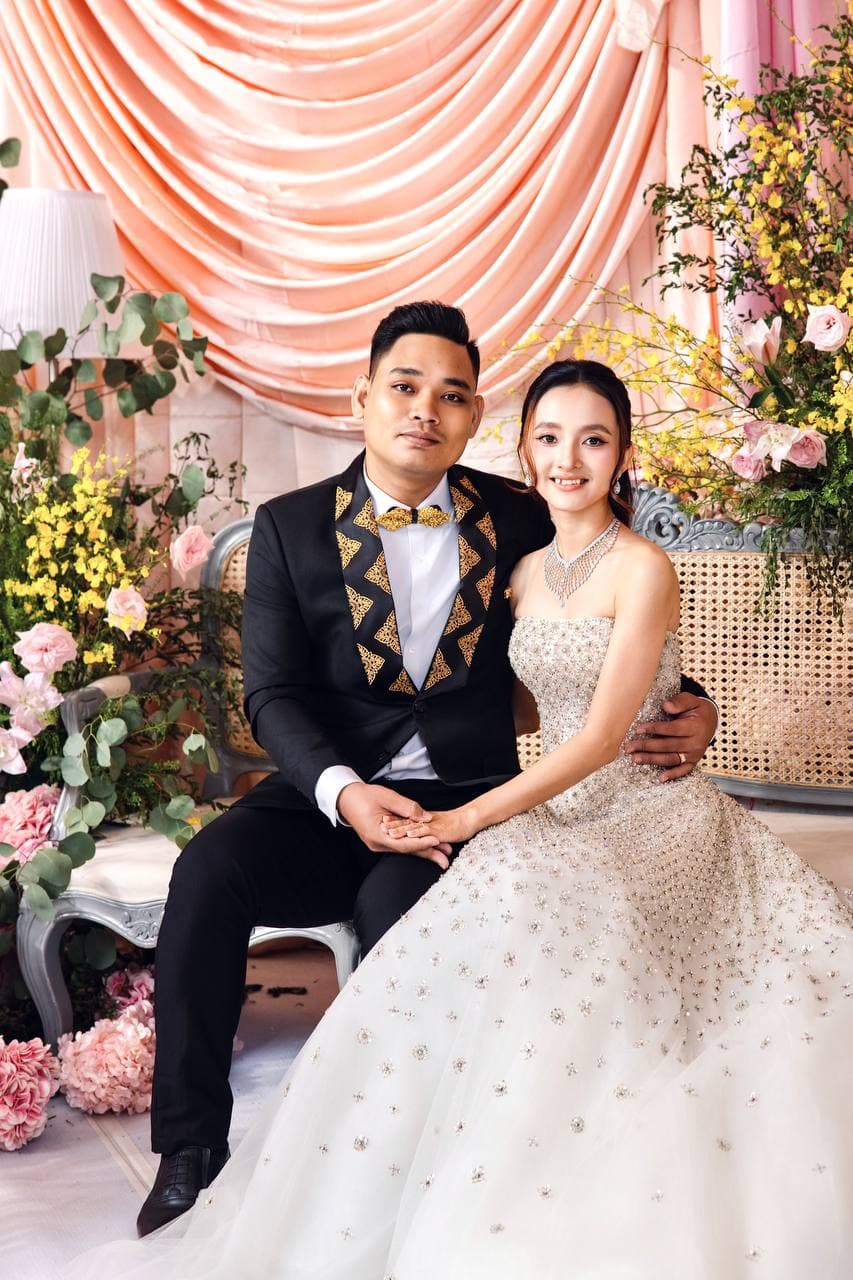 Sokvebol & Souhouy — គូស្វាមីភរិយាអង្គុយក្បែរគ្នាក្នុងចំណោមផ្កាពណ៌ផ្កាឈូក និងលឿង ក្នុងអាវពាក់ស៊ុមមាស និងរ៉ូបពណ៌សប៉ាក់គុជ